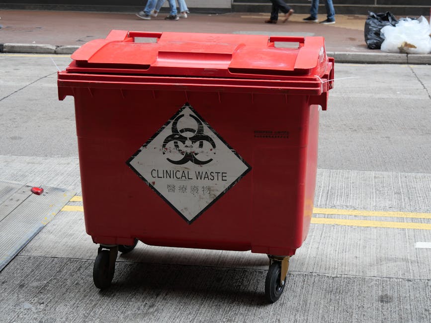 A red wheeled bin labeled with a biohazard symbol and the words 'Clinical Waste' is positioned on a concrete sidewalk outside a building, with a group of pedestrians walking along the street in the background. The bin has a hinged lid, and the label indicates it is used for disposal of medical or hazardous waste. Surrounding the bin, there are small yellow traffic lines painted on the pavement and some scattered debris. Nearby, there are black rubbish bags placed on the pavement, and an individual is seen in the background. This scene may be part of a house removal or relocation process involving the transfer of specialized waste materials, as coordinated by services like Man with Van Chase Cross, ensuring safe logistics and proper disposal during home moves.