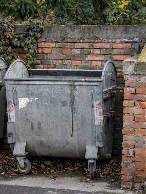 A red wheeled bin labeled with a biohazard symbol and the words 'Clinical Waste' is positioned on a concrete sidewalk outside a building, with a group of pedestrians walking along the street in the background. The bin has a hinged lid, and the label indicates it is used for disposal of medical or hazardous waste. Surrounding the bin, there are small yellow traffic lines painted on the pavement and some scattered debris. Nearby, there are black rubbish bags placed on the pavement, and an individual is seen in the background. This scene may be part of a house removal or relocation process involving the transfer of specialized waste materials, as coordinated by services like Man with Van Chase Cross, ensuring safe logistics and proper disposal during home moves.