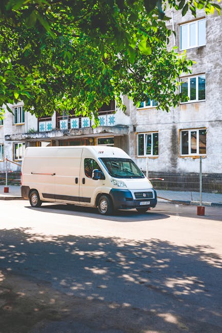 A white cargo van from Man with Van Chase Cross is parked on a residential street with a multi-storey apartment building in the background. The van is positioned parallel to the pavement, with its fro
