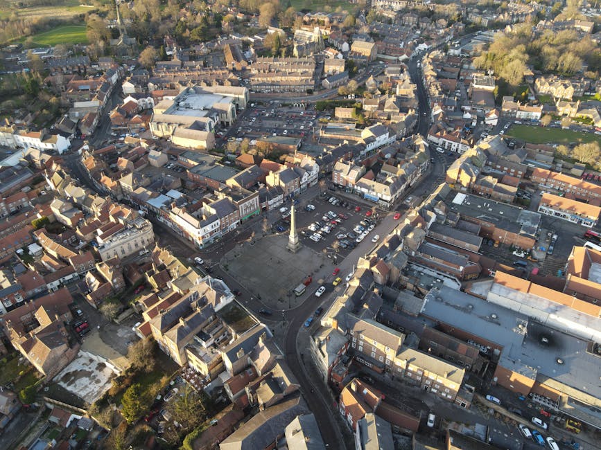 An aerial view of a town centre in Chase Cross showing tightly packed residential and commercial buildings with narrow streets and a central town square featuring a tall monument. The square is surrounded by parked cars, with a few vehicles moving along the roads. The buildings include brick terraced houses, small shops, and some modern structures. In the foreground, rooftops and backyards are visible, while the background reveals more greenery and mature trees. The scene is illuminated by natural daylight, highlighting the details of the urban environment. This image illustrates the urban setting where Man with Van Chase Cross provides house removals and relocation services, including furniture transport and packing for home moves within the local area.