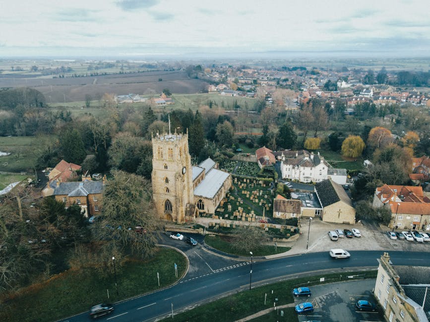 Aerial view of a small village showing a historic stone church with a tower and arched windows, surrounded by residential houses with red and grey tiled roofs. In front of the church, a graveyard with numerous headstones and well-maintained green hedges is visible. A narrow paved road curves past the church, with several cars parked along the side and a few driving by. To the left of the church, large leafy trees partially obscure some residential buildings. The background reveals open fields, patchworks of farmland, and distant countryside under a cloudy sky. The scene captures a peaceful, rural environment, illustrating the kind of setting where household relocation services, such as those offered by Man with Van Chase Cross, might assist with home removals and furniture transport in a traditional village context.