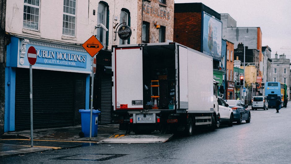 A white box truck with an open back door is parked on a street in front of a row of commercial buildings, including Dublin Mouldings with a blue sign. The truck is positioned partially on the curb wit