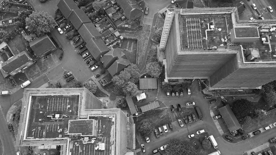 Aerial view of a residential area showing a multi-storey apartment building with a flat roof equipped with solar panels, surrounded by smaller houses, trees, and a mixture of parked cars along the streets and in a designated parking area. In the foreground, a man with a van from Man with Van Chase Cross is seen parked on the pavement near the entrance to the apartment complex. The van's side door is open, revealing packing materials such as cardboard boxes, plastic-wrapped furniture, and blankets used for home relocation. A move is in progress with a person wearing casual clothing actively loading or unloading the boxes and furniture from the van onto the pavement or into the building doorway. The scene captures the logistical aspects of flexible furniture transport and packing and moving operations typical of house removals services on the Collier Row Estate to Chase Cross, with the surrounding environment including greenery, residential dwellings, and parking facilities all visible from the aerial perspective.