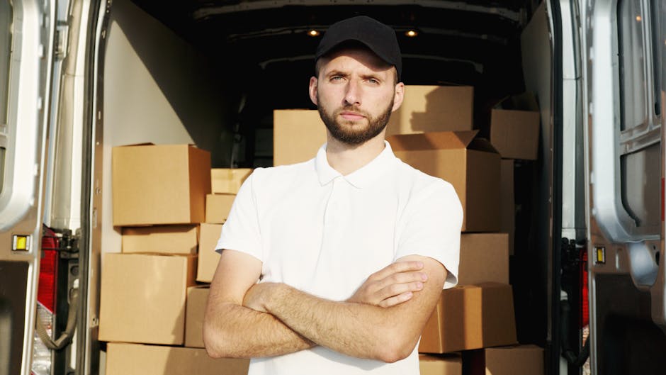 A man wearing a blue beanie, red and navy jacket, and black trousers stands with arms crossed in front of an open van with its rear doors flung wide. The van is loaded with neatly stacked cardboard bo
