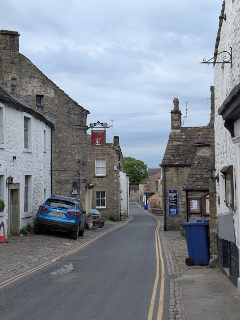 A narrow residential street in Chase Cross featuring older stone and brick houses with slate roofs and small windows. A blue car is parked on a cobblestone driveway adjacent to a white-painted house on the left side, with a small bench nearby. On the right side, there is a blue wheeled rubbish bin placed on the sidewalk next to the outer wall of a building. The street is paved with asphalt and has double yellow lines along the curb, indicating parking restrictions. The scene is captured during daylight with a mostly cloudy sky overhead, and a red signboard hanging from a metal bracket on one of the buildings in the background. The overall setting suggests a quiet, historic neighbourhood potentially involved in house removals or home relocation activities, with a focus on transport and logistics for moving furniture and belongings.