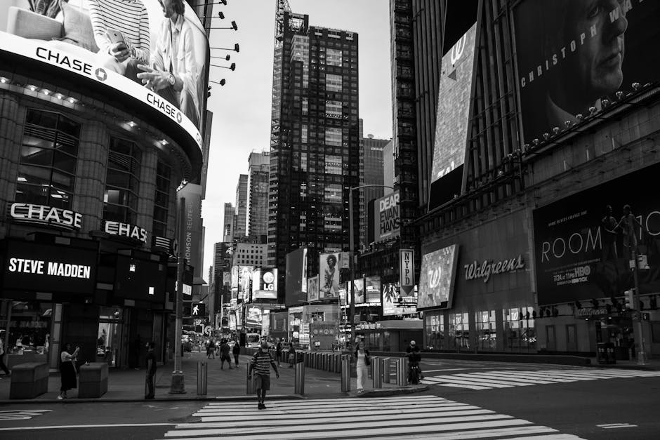 Black and white photograph of a busy urban city centre with tall commercial buildings and billboards, including prominent signage for 'Chase' and 'Steve Madden.' Pedestrians are walking across a crosswalk on the pavement, some carrying shopping bags or backpacks. The scene shows several storefronts, advertising screens, and digital billboards on the buildings, with a high density of structures typical of a city street. The environment appears lively, with people engaged in daily activities, and the area is illuminated by ambient city lighting. The image captures the essence of street-by-street movement logistics that companies like Man with Van Chase Cross accommodate when offering house removals and furniture transport services amidst urban environments.