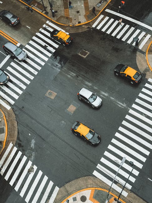 An aerial view of a city street intersection with four pedestrian crossings marked by white striped lines. Several cars, including two yellow taxis, a silver vehicle, and a darker car, are driving through the intersection on a wet asphalt surface. The surrounding pavement features curbs, a small roundabout with a patterned design, and a few pedestrians waiting to cross. The scene appears to be during daytime with overcast weather, and the environment suggests an urban area, possibly near residential or commercial buildings. This image may be used to support content about urban house removals or moving logistics, highlighting the challenges of navigating city streets during a home relocation or furniture transport process as part of a professional removal service like Man with Van Chase Cross.