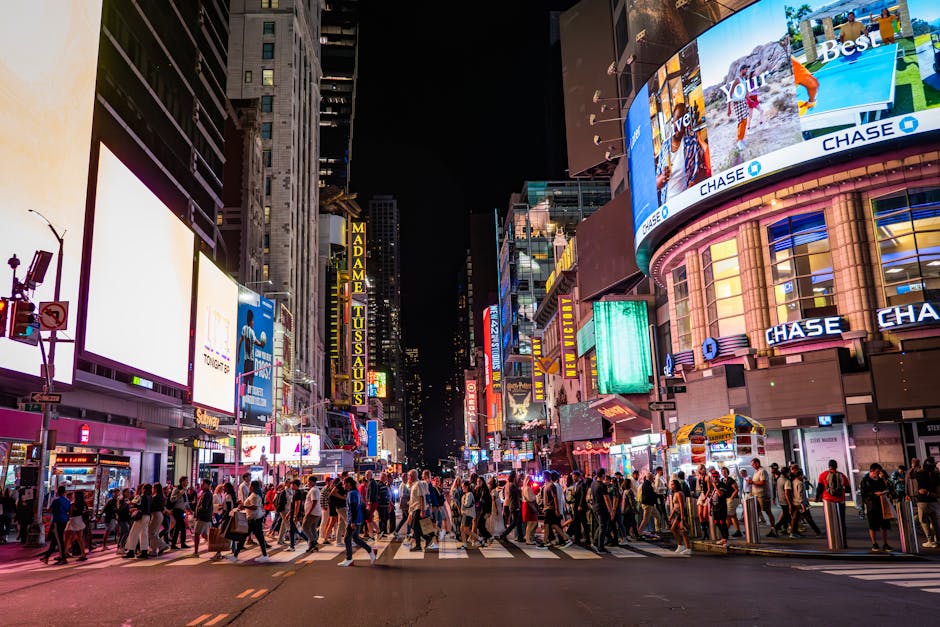 Black and white photograph of a busy urban city centre with tall commercial buildings and billboards, including prominent signage for 'Chase' and 'Steve Madden.' Pedestrians are walking across a crosswalk on the pavement, some carrying shopping bags or backpacks. The scene shows several storefronts, advertising screens, and digital billboards on the buildings, with a high density of structures typical of a city street. The environment appears lively, with people engaged in daily activities, and the area is illuminated by ambient city lighting. The image captures the essence of street-by-street movement logistics that companies like Man with Van Chase Cross accommodate when offering house removals and furniture transport services amidst urban environments.