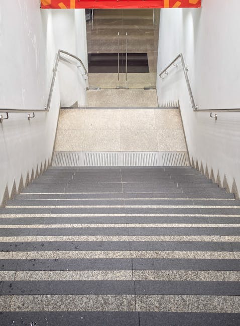 Inside a building, a set of grey carpeted stairs with black anti-slip strips on each step descends towards a doorway equipped with metal handrails on both sides. Beyond the stairs, a tiled area with a black doormat is visible, leading to an open glass door. The doorway provides access to an outdoor pavement area. The image captures the interior part of a home relocation process, with the stairs presenting a potential access challenge for moving large furniture or boxed belongings during a house move. The lighting is bright, suggesting daytime, and the environment appears clean and well-maintained. Man with Van Chase Cross may be involved in the furniture transport and packing and moving services, as implied by the scene.