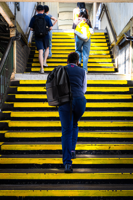 A man with a black backpack is ascending a set of outdoor stairs with yellow and black painted steps, leading to an underpass or entrance illuminated by natural daylight. At the top of the stairs, three individuals—two men and one woman—are standing and engaged in conversation or preparing to move. The woman is wearing a yellow jacket and holding a bag, while the men are dressed casually, one in a black cap and backpack, the other in a dark shirt. The scene is part of a home relocation process, with the stairs potentially posing access challenges, which Man with Van Chase Cross addresses through their removals services. The environment appears to be urban, with concrete walls and metal railings visible along the staircase, and the overall setting suggests a busy residential or transport hub. The image captures the active loading or transport phase of a furniture or household items move, emphasizing careful navigation of access points during a house removal in RM6.