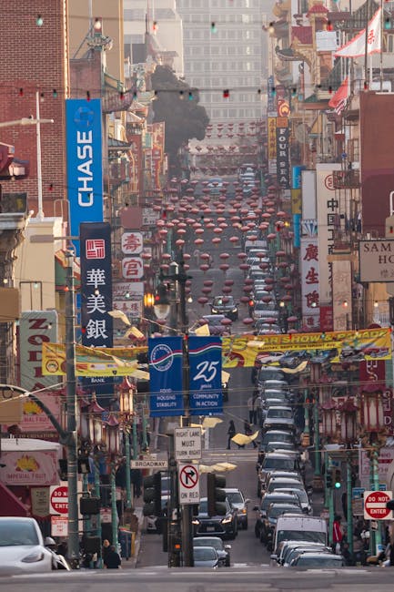 A busy urban street scene in Chinatown, with numerous parked cars lining both sides of the road and traffic lights at the intersection. The street is densely packed with various storefronts displaying colorful signs in Chinese and English, advertising different businesses, including a bank and Asian supermarkets. Above the street, strings of red lanterns are suspended, creating a festive atmosphere. Smoke is visible rising from a grill or food stall on the sidewalk, indicating outdoor dining or street food activity. In the background, tall modern buildings and a smoky haze are seen, suggesting ongoing urban activity. The street is illuminated by natural daylight and traffic signals, providing a clear view of the lively environment. This scene relates to house removals and relocation services as part of moving logistics in a busy city setting, with Man with Van Chase Cross potentially assisting with transportation and packing during a home relocation process.