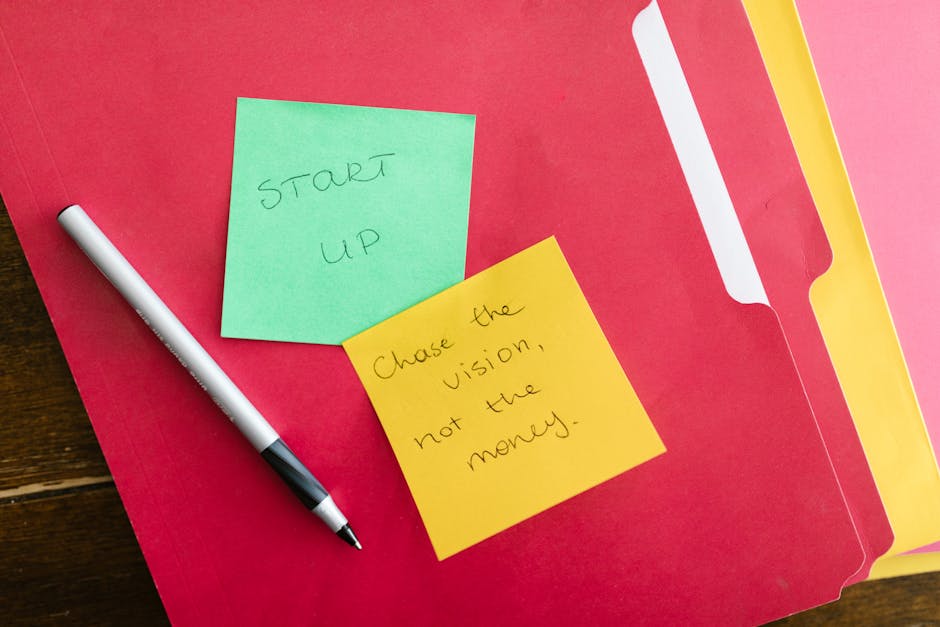 A red file folder is open on a wooden surface with three yellow and green sticky notes placed on top. One green sticky note has handwritten text that reads 'START UP,' while a yellow sticky note below it contains the message 'Chase the vision, not the money.' A black and white marker lies diagonally across the folder, suggesting recent writing or planning. The scene relates to organizing or preparing for home relocation, with the sticky notes symbolising key ideas or motivational reminders associated with moving services, as offered by Man with Van Chase Cross, and connects to the topic of understanding removals pricing and accurate quotes for house removals.